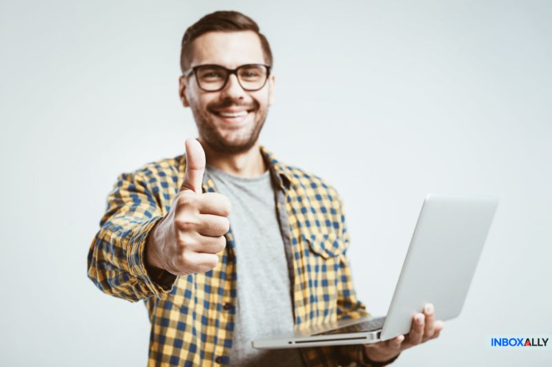 A man holding a laptop gives a thumbs up and smiles at the camera, possibly relieved after resolving email errors such as 550 5.7.0 Local Policy Violation. He is wearing glasses and a yellow plaid shirt against a plain, light-colored background.