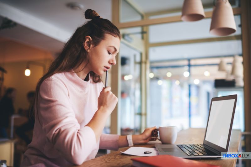 A woman sits at a table in a cafe, looking at her laptop screen while holding a pencil to her mouth, trying to solve a 550 5.7.0 error related to email size limit, with a notebook and coffee mug nearby.