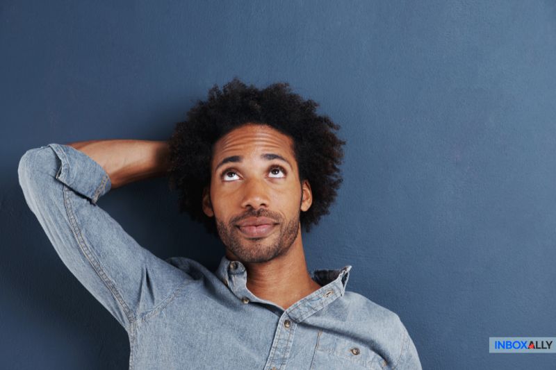 Un hombre con camisa vaquera está de pie contra una pared azul, mirando hacia arriba con una mano apoyada en la nuca, como si estuviera reflexionando sobre el mejor servicio de correo electrónico, como Mailgun o Amazon SES, para las necesidades de su empresa.