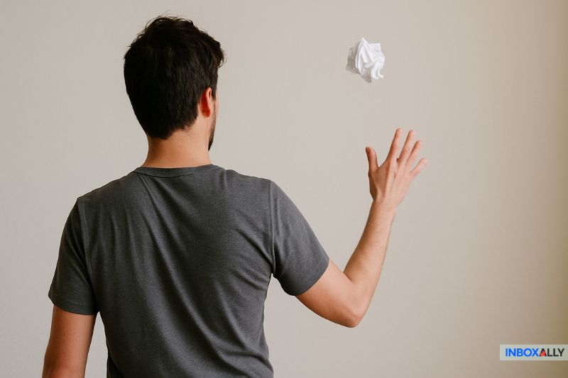 A man in a gray t-shirt tosses a crumpled piece of paper toward a wall, symbolizing the hassle of using a disposable email address. The logo "INBOXALLY" appears in the lower right corner.