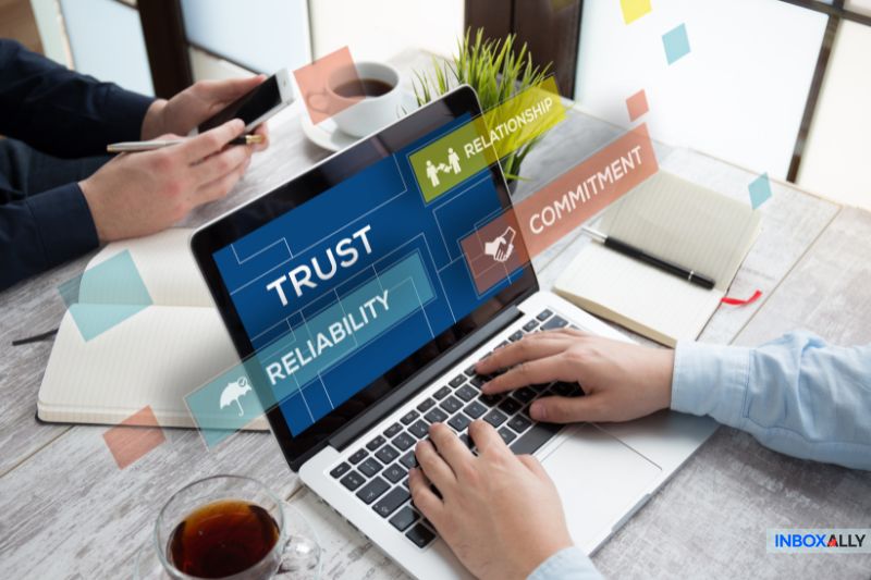 Two people work at a desk with a laptop displaying infographic keywords: Trust, Relationship, Commitment, Reliability, and the benefits of using a disposable email address. Notebooks, coffee, and a phone are also on the desk.
