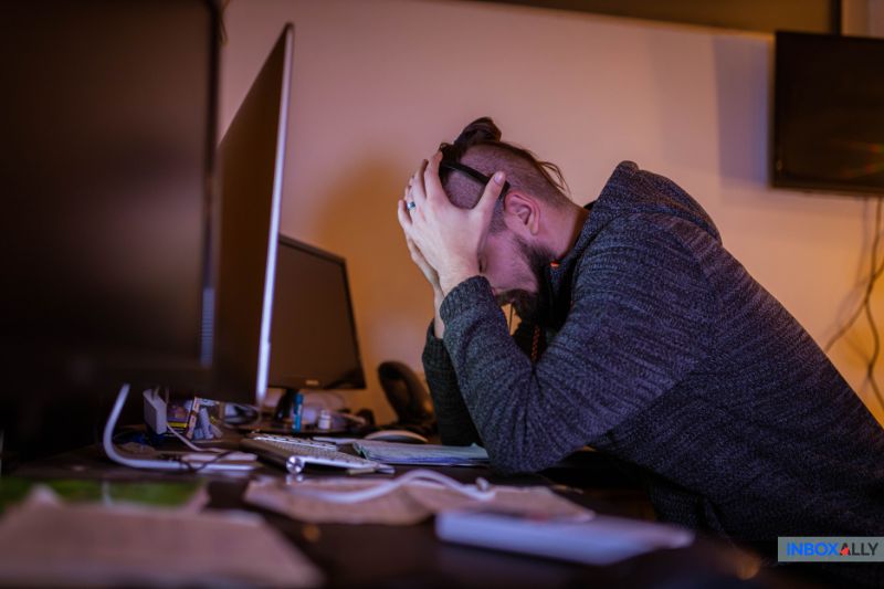 A person sits at a desk with their head in their hands, appearing stressed or frustrated in front of a computer monitor, possibly overwhelmed by managing multiple temporary email accounts.